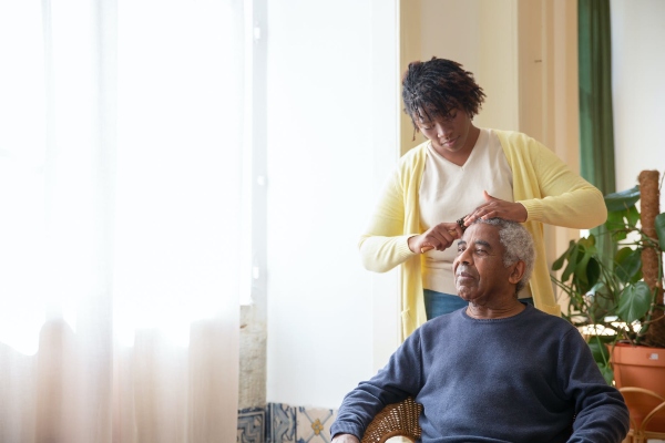An elderly man with dementia stares out the window while his caregiver helps to comb his hair.