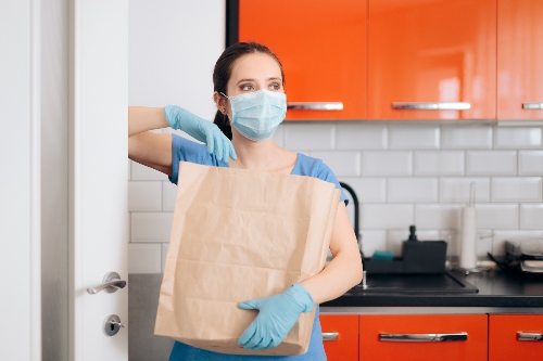 A home caregiver wears a protective face mask, gloves, and scrubs while helping to carry a grocery bag.